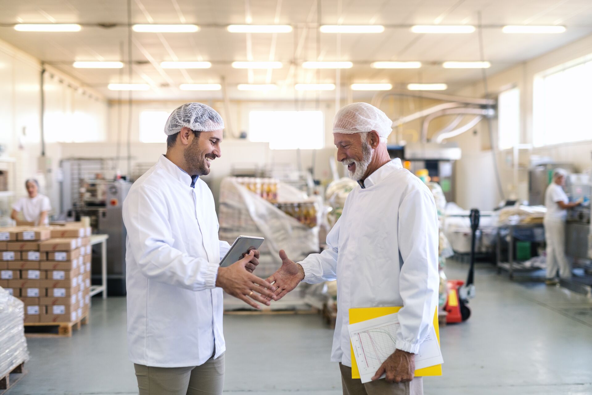 Two businessmen in sterile uniforms shaking hands for good done job while standing in food factory. Older one holding folder and graphs while younger holding tablet