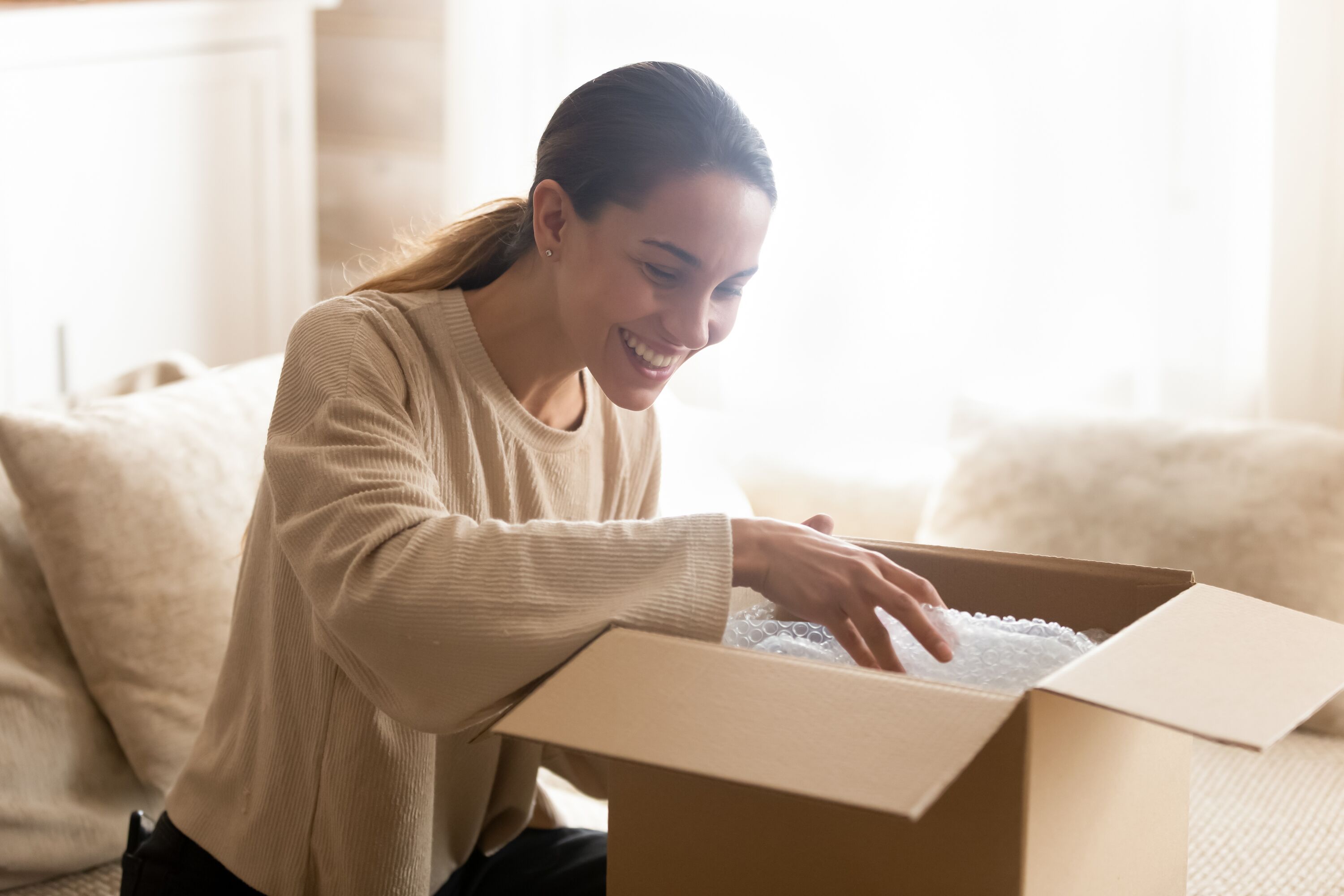 Smiling millennial woman sit on couch at home open post package shopping online buying goods on internet, happy young female customer unpack postal shipping parcel satisfied with order or delivery