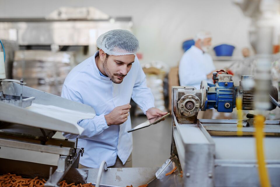 Young Caucasian serious supervisor evaluating quality of food in food plant while holding tablet. Man is dressed in white uniform and having hair net