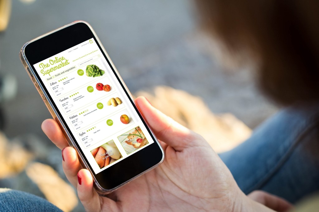 close-up view of young woman shopping groceries on online supermarket with her mobile phone. All screen graphics are made up.