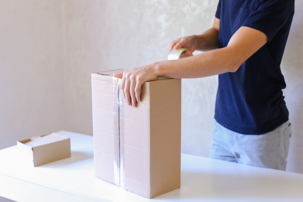 Deliveryman Standing in Room, Glue Tape Box and Takes Parcel in Hands and Smiling . Postman European Appearance in a Black T-Shirt and Cap Stands Near the Desk in the Office With White Walls and Unassisted Packaging Boxes, Adhesive Glue Tape on a Cardboard Box, the Courier Turns Box Vertically, Taking the Box in His Hands and Smiling at the Camera. Concept of Work in Office and Favorite Work 24 Hours Day, Mail Delivery From Post Office,free Delivery Worldwide or Delivery on Time, Advertising of New Shipping Service Company, Purchase at Any Time and Any Day.