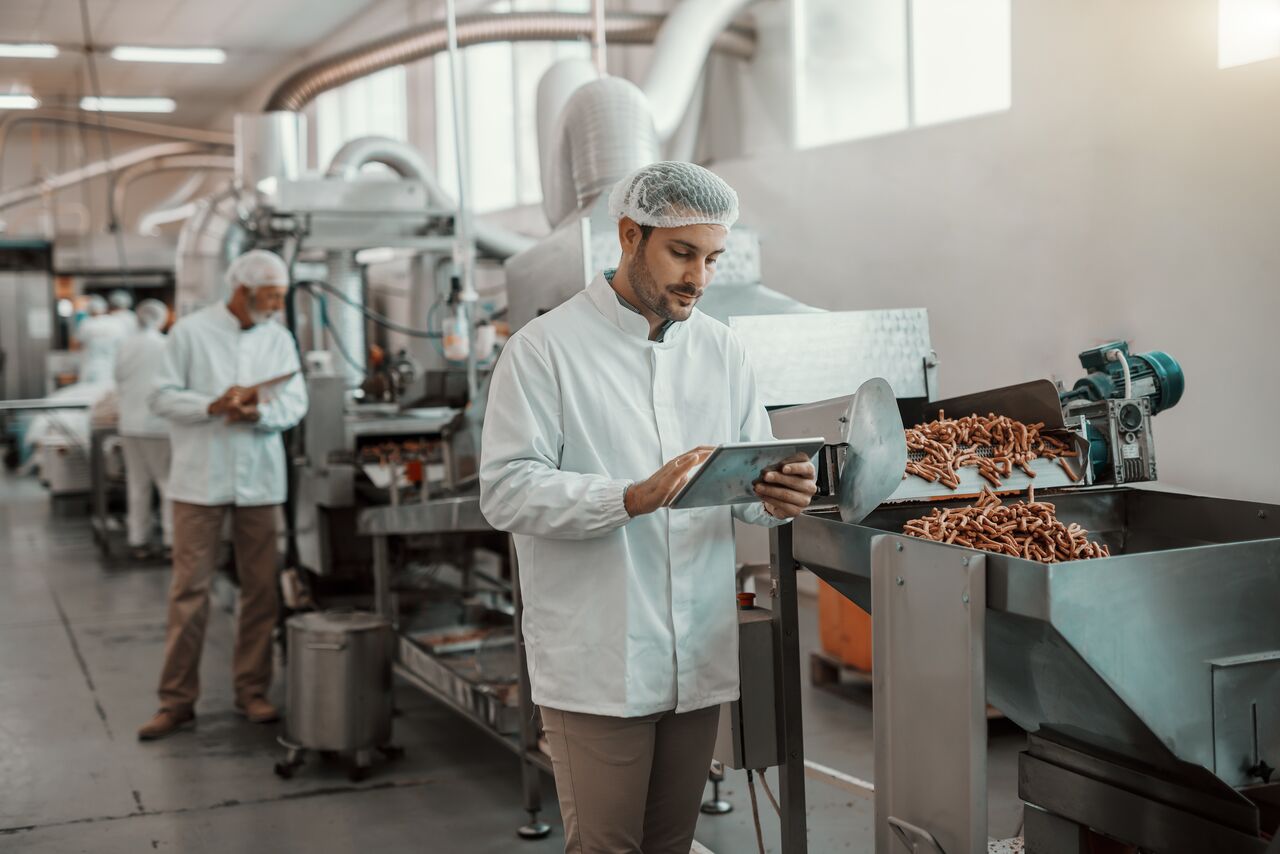 Young Caucasian serious supervisor evaluating quality of food in food plant while holding tablet. Man is dressed in white uniform and having hair net